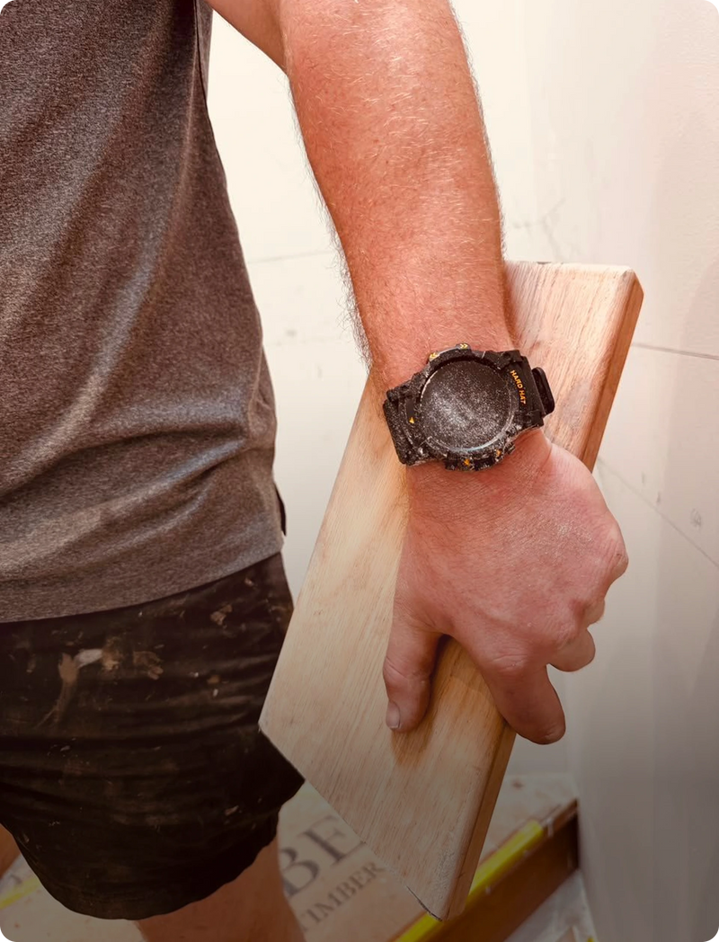 A person wearing a rugged black digital watch with a textured, distressed dial, holding a light-colored wooden board in a workshop setting.