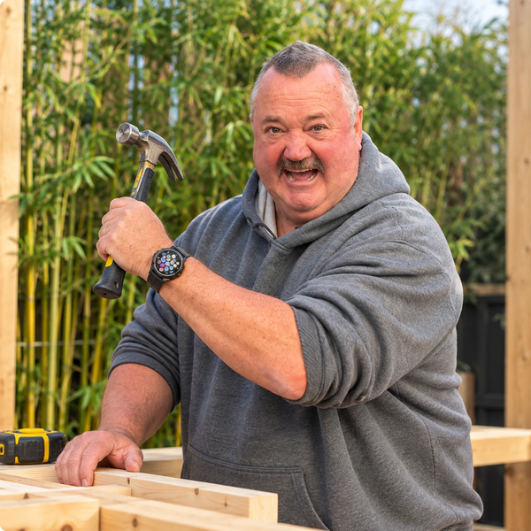 A smiling man wearing a gray hoodie holds a hammer while working on a wooden structure outdoors, with bamboo fencing in the background.